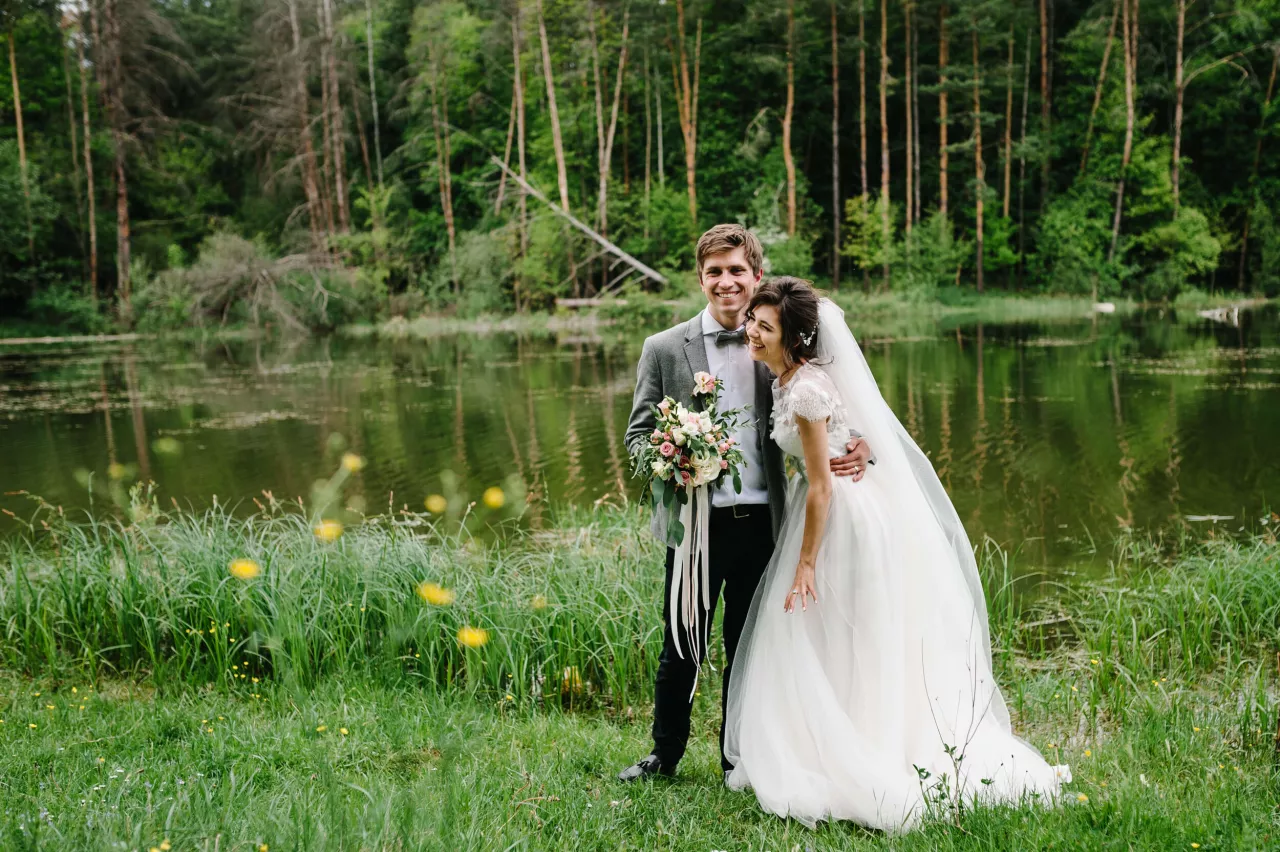 Bride and groom smiling in forest with water behind them